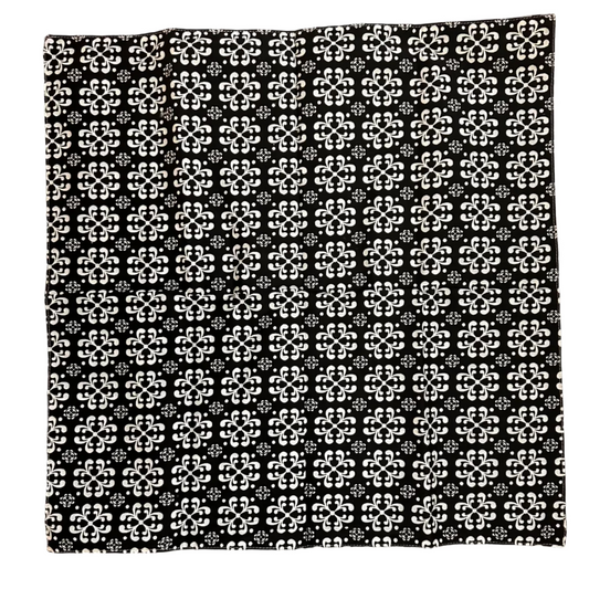 A square black fabric item, a napkin, featuring a repeating white geometric floral pattern, laid flat on a brown wooden floor.