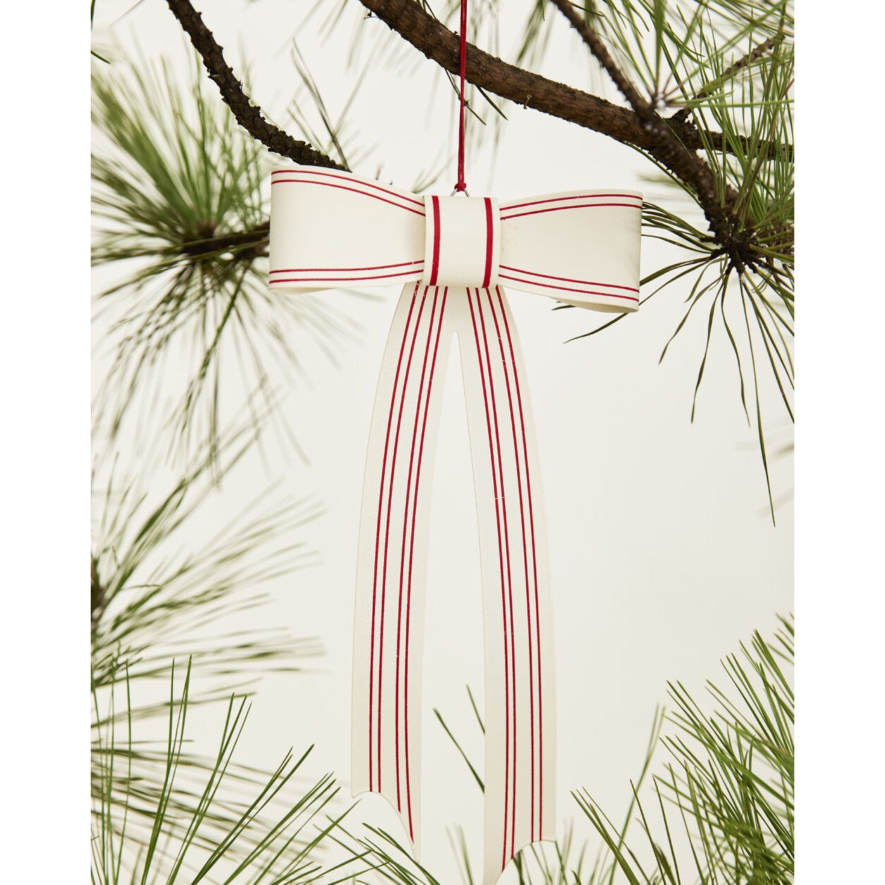 A white metal Christmas ornament shaped like a bow with red stripes, hanging from a pine branch.