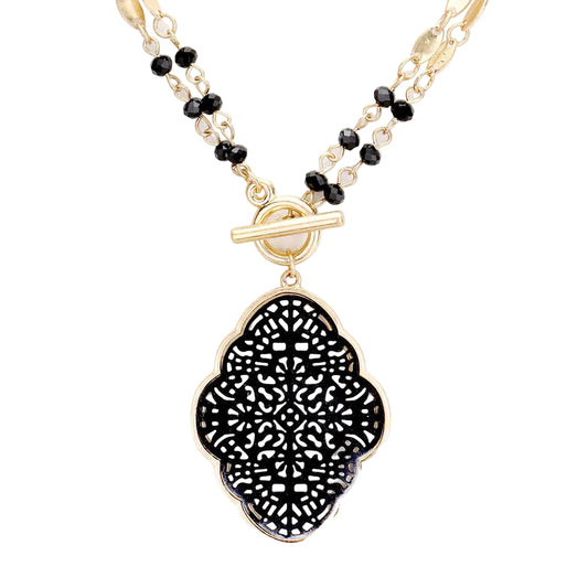 Gold-tone necklace featuring a large black filigree clover pendant, a toggle clasp, and a beaded chain with black faceted beads and clear crystal accents, shown on a solid white background.