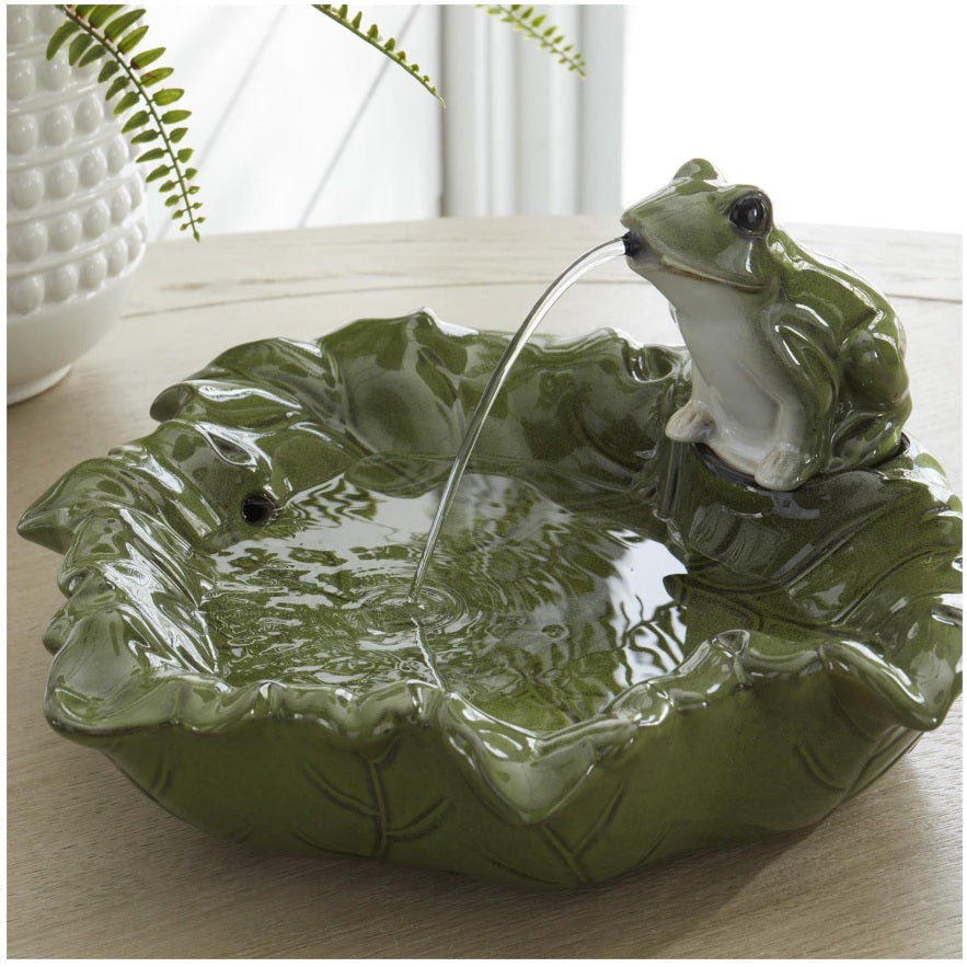 A green ceramic frog fountain bowl with a glossy finish, featuring a small frog perched on the rim from which water flows into a leaf-shaped basin, set on a light wooden surface.