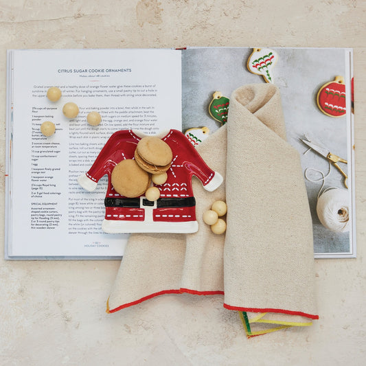 A flat-lay of a Christmas-themed cookie ornament set on an open cookbook. The set includes a red and white ceramic Santa cookie ornament, several beige wooden beads, and two cream-colored felted wool cookie cutters with red trim, styled on a light beige surface.