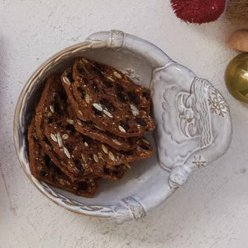 A ceramic bowl with a Santa Claus face design on the side, filled with dark chocolate chip cookies or pretzels on a light textured surface.