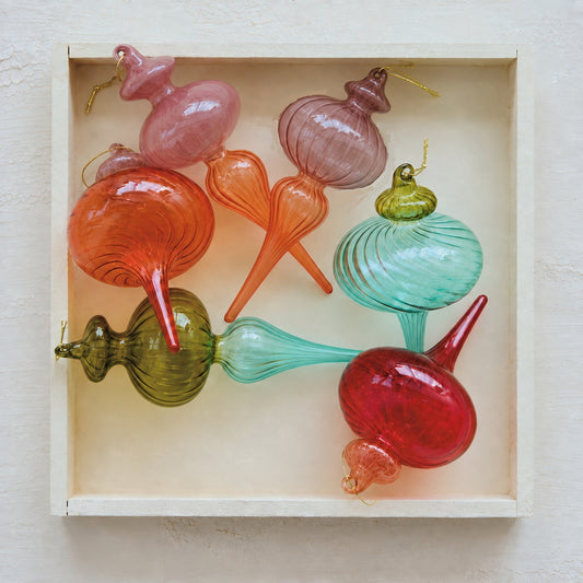 A set of colorful hand-blown glass ornaments displayed inside a square white wooden box against a light beige textured wall.