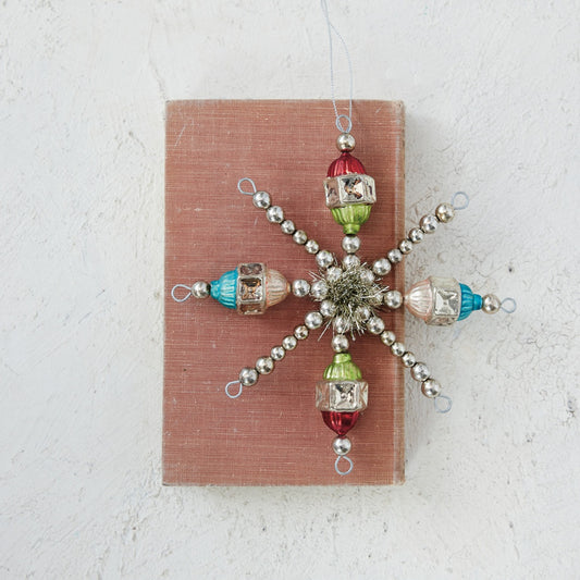 A vintage-style snowflake ornament made of silver beads and colorful glass beads, displayed on a dusty pink rectangular card against a textured off-white background.