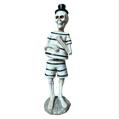 A decorative skeleton figurine dressed in a black-and-white striped sailor shirt and shorts, wearing a small black top hat and standing on a round base against a plain white background.