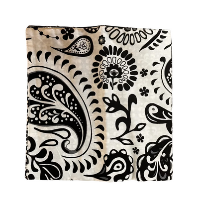 A square fabric item, a napkin, featuring a black paisley and floral block print on a cream-colored background, laid flat on a white surface