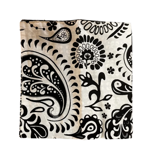 A square fabric item, a napkin, featuring a black paisley and floral block print on a cream-colored background, laid flat on a white surface