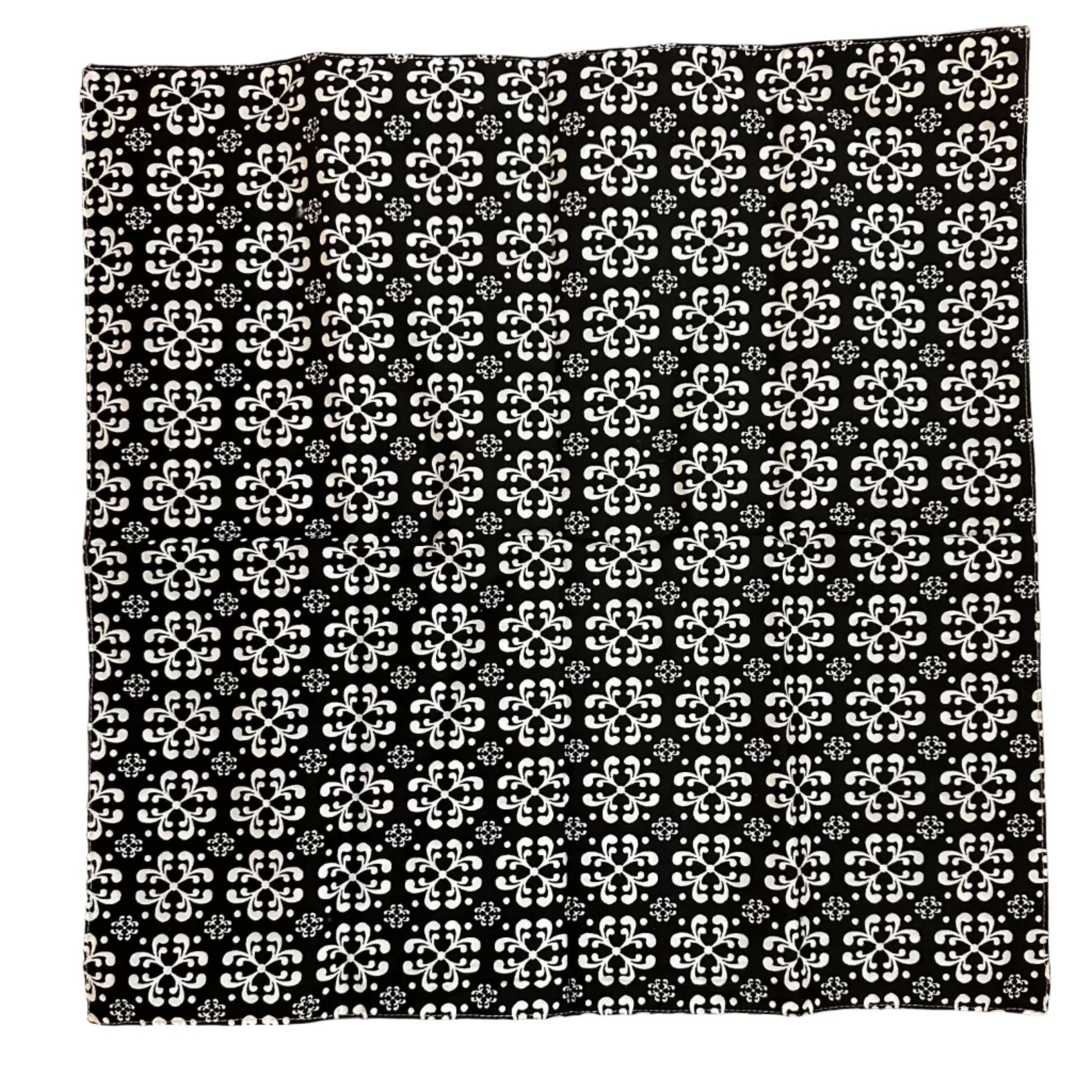 A square black fabric item, a napkin, featuring a repeating white geometric floral pattern, laid flat on a brown wooden floor.