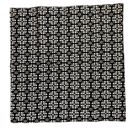 A square black fabric item, a napkin, featuring a repeating white geometric floral pattern, laid flat on a brown wooden floor.