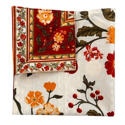 A folded floral fabric napkin with a cream background and orange, red, and green botanical print, displayed on a white background.