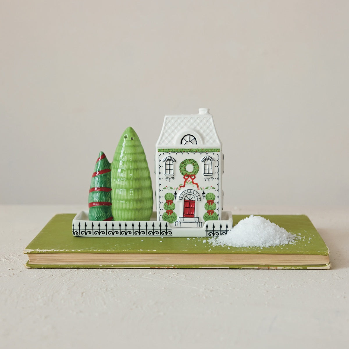 A small holiday scene set on a book, featuring salty and pepper shakers:  white house with green wreath and red door, two green trees, and a pile of salt (white faux snow) on a beige surface against a light neutral background.