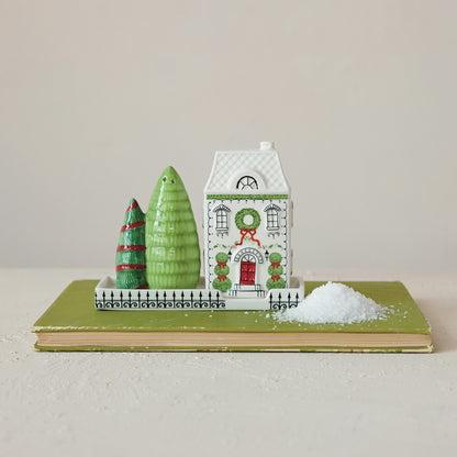 A small holiday scene set on a book, featuring salty and pepper shakers:  white house with green wreath and red door, two green trees, and a pile of salt (white faux snow) on a beige surface against a light neutral background.