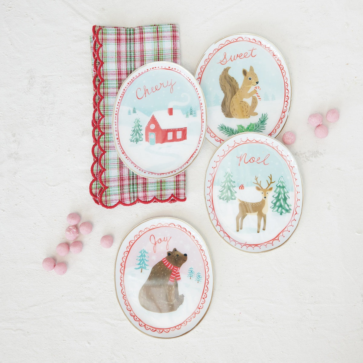 Four oval Christmas-themed dish trays with illustrated woodland animals and words like 'Cherry,' 'Sweet,' 'Noel,' and 'Joy,' arranged on a white textured surface beside a red-and-green plaid napkin and pink candy decorations.