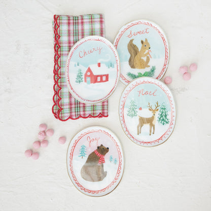 Four oval Christmas-themed dish trays with illustrated woodland animals and words like 'Cherry,' 'Sweet,' 'Noel,' and 'Joy,' arranged on a white textured surface beside a red-and-green plaid napkin and pink candy decorations.