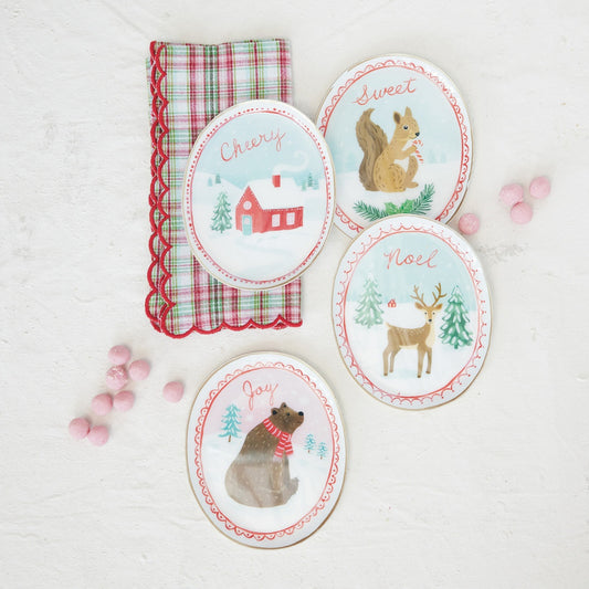 Four oval Christmas-themed dish trays with illustrated woodland animals and words like 'Cherry,' 'Sweet,' 'Noel,' and 'Joy,' arranged on a white textured surface beside a red-and-green plaid napkin and pink candy decorations.