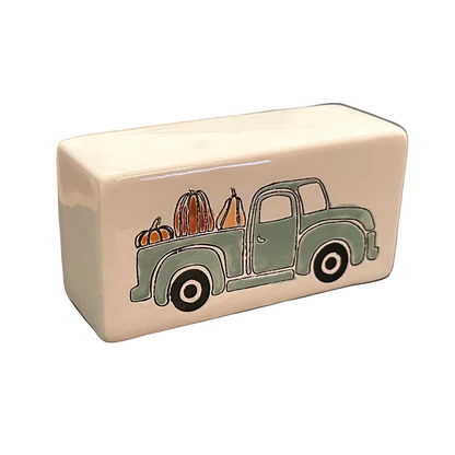 A white rectangular block with a vintage green pickup truck carrying pumpkins, resting on a black laptop on a wooden kitchen table.