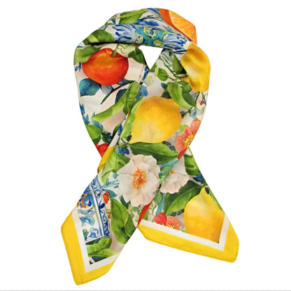 A square silk scarf with a vibrant citrus and floral print, styled in a loop over a white gift box. The print features yellow lemons, red oranges, white flowers, and green leaves on a cream background with blue patterned borders.