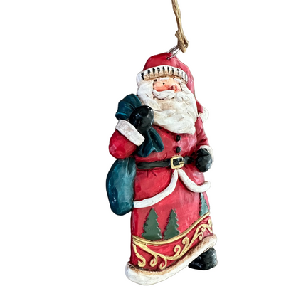 A hand-painted Christmas hanging ornament shaped like Santa Claus, featuring a red coat with white fur trim, a dark green sack, and gold accents, hanging on a white surface.