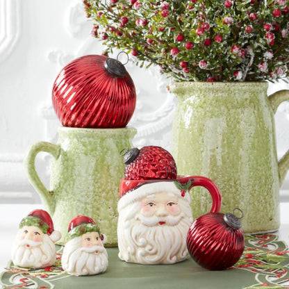 A festive holiday tabletop arrangement featuring a ceramic Santa mug with a red textured hat, two small Santa figurines, a large red ribbed ornament resting on a green mug, and another red ornament beside the mug, with a green pitcher holding red berries in the background.