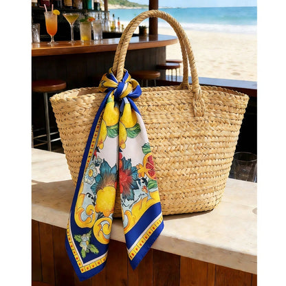 A woven straw tote bag with two top handles sits on a beach bar counter, with a colorful printed scarf tied around the handle. The background shows a blurred beach and ocean scene.