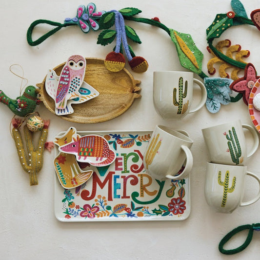 A collection of handmade Christmas-themed felt crafts and decorations arranged on a light beige background, including a green felt garland with hearts, flowers, and butterflies, a pink felt nativity scene oval tray, a white felt 'Merry Christmas' rectangular tray, three white ceramic mugs with green cactus prints, a small felt bird ornament, and a gold felt angel decoration.
