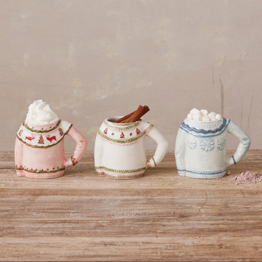 Three ceramic mug-shaped containers filled with whipped cream, arranged on a wooden surface against a textured beige wall. The left mug is light pink with red holiday motifs, the center mug is cream with green trim and a cinnamon stick, and the right mug is white with blue trim and snowflake details.