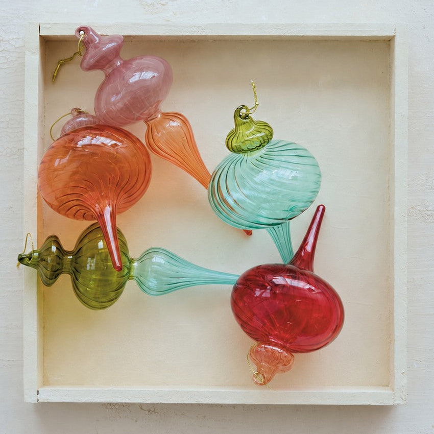 A set of colorful hand-blown glass ornaments shaped like stylized bulbs and teardrops, arranged inside a small light wood box on a textured off-white background.