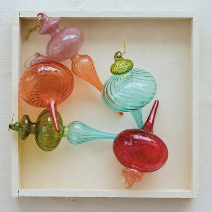 A set of colorful hand-blown glass ornaments shaped like stylized bulbs and teardrops, arranged inside a small light wood box on a textured off-white background.
