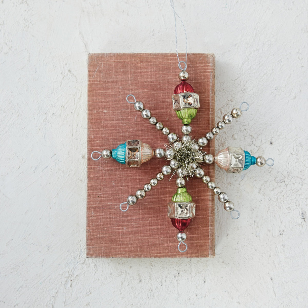 A vintage-style snowflake ornament made of silver beads and colorful glass beads, displayed on a dusty pink rectangular card against a textured off-white background.