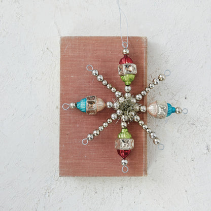 A vintage-style snowflake ornament made of silver beads and colorful glass beads, displayed on a dusty pink rectangular card against a textured off-white background.