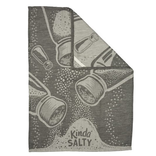 A square gray textile item, a towel, featuring a printed pattern of tilted salt and pepper shakers and the words 'Kinda SALTY' in a playful font. One corner is folded over to show the reverse side, which is a light gray textured fabric.